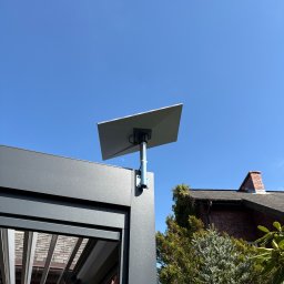 Systemy Zabezpieczen Satklinika - Nowoczesna antena satelitarna zamontowana na pergoli, na tle błękitnego nieba i fragmentu domu z cegły. Widoczne elementy konstrukcyjne pergoli.