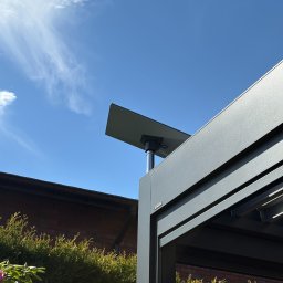 Systemy Zabezpieczen Satklinika - Nowoczesna pergola z panelem słonecznym na tle błękitnego nieba i zieleni. Minimalistyczny design, szara konstrukcja, widok z dołu.