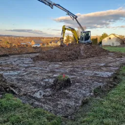 Żółta mini koparka na placu budowy, przygotowująca teren pod fundamenty; widoczny wykop, urobiona ziemia i ramię pompy do betonu na tle zabudowań mieszkalnych i błękitnego nieba.