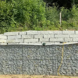 Gabionowa konstrukcja oporowa z warstwą wyrównującą z jasnych, prostokątnych bloków kamiennych, widoczny żółty przewód na tle zieleni.