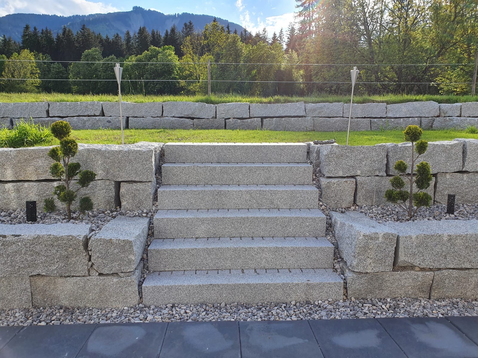 Granitowe schody zewnętrzne z czterema stopniami, otoczone kamiennym murem i ozdobnymi krzewami, z widokiem na zielony trawnik, drzewa i góry w tle.