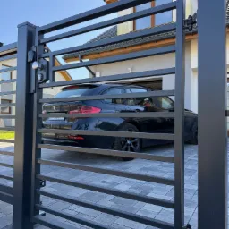 Nowoczesna, pozioma brama wjazdowa w kolorze antracytowym, z widocznym samochodem na posesji. Brama z zamkiem i klamką, na tle domu z jasną elewacją i grafitową dachówką.