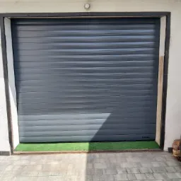 Zamknięta, szara brama garażowa segmentowa z poziomymi panelami, zamontowana w białej elewacji z ciemnym obramowaniem, z fragmentem sztucznej trawy u dołu.