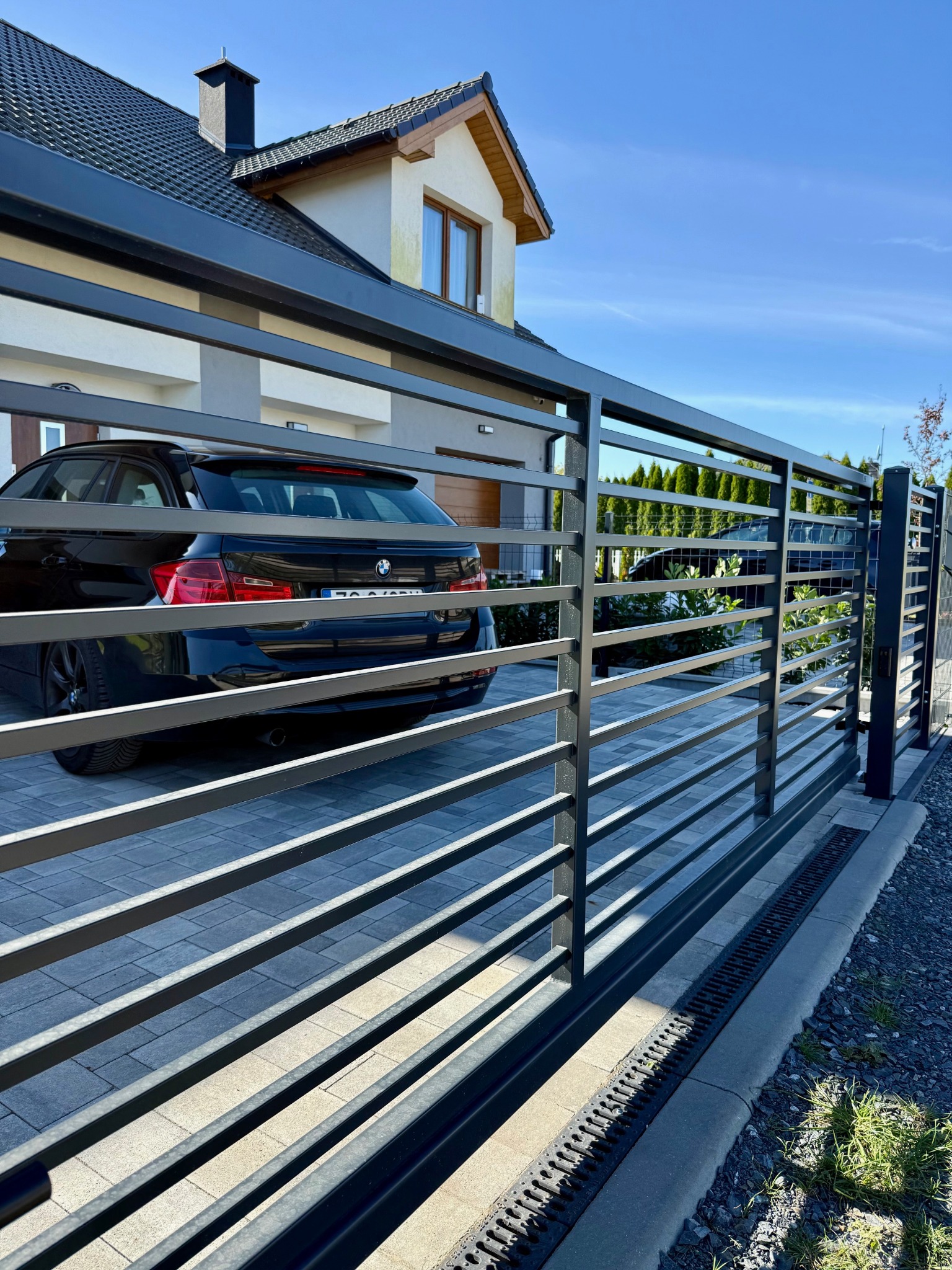 Nowoczesna, pozioma brama wjazdowa z metalowymi profilami, na tle domu z czarnym samochodem. Brama w kolorze grafitowym, z widoczną kostką brukową i zielenią w tle.