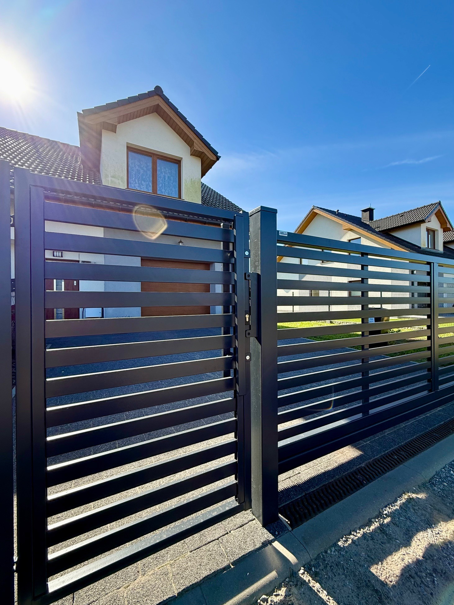 Nowoczesna, czarna brama wjazdowa z poziomymi panelami na tle domu z beżową elewacją i błękitnego nieba w słoneczny dzień. Widoczny fragment chodnika.