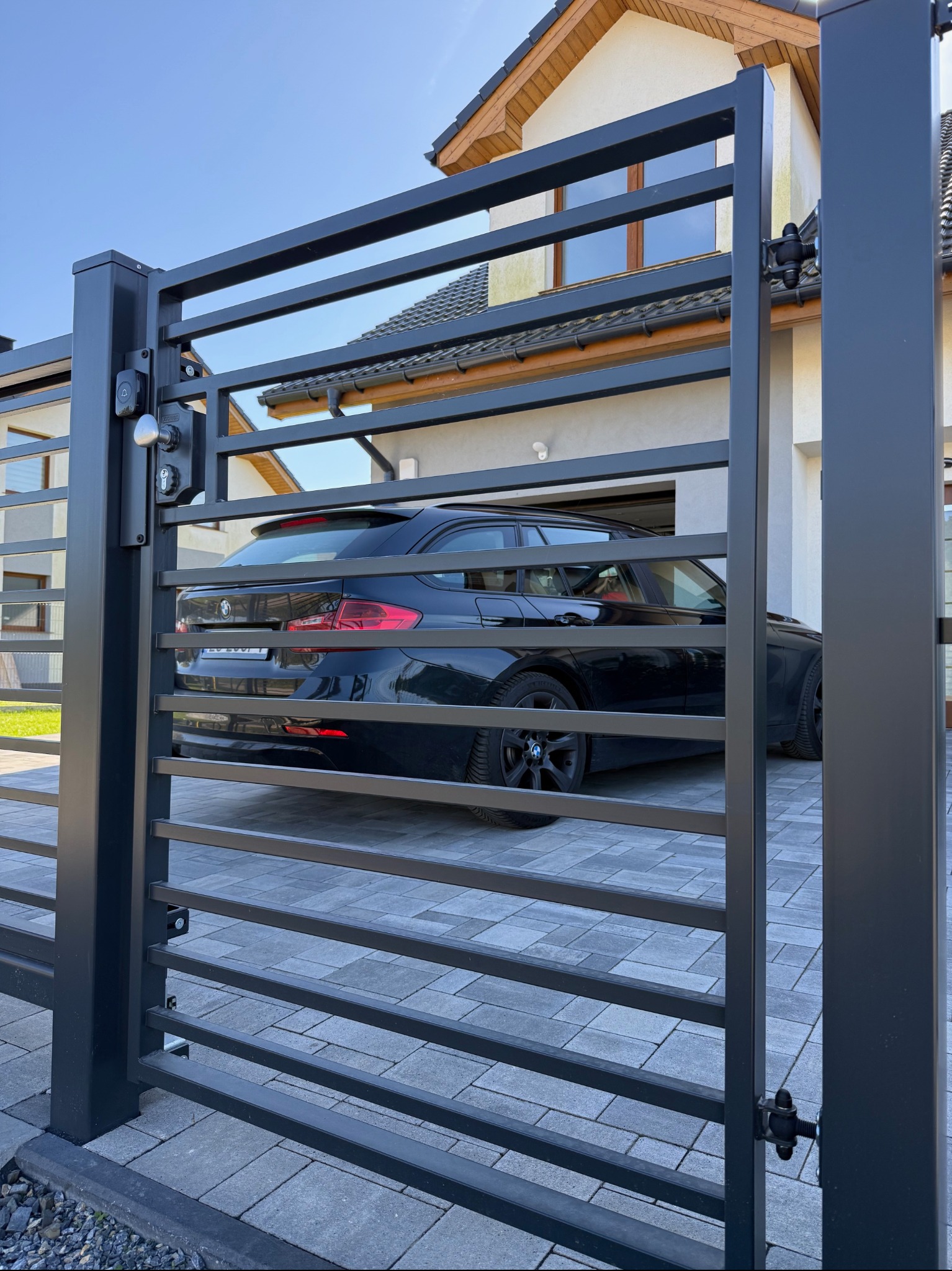 Nowoczesna, pozioma brama wjazdowa w kolorze antracytowym, z widocznym samochodem na posesji. Brama z zamkiem i klamką, na tle domu z jasną elewacją i grafitową dachówką.