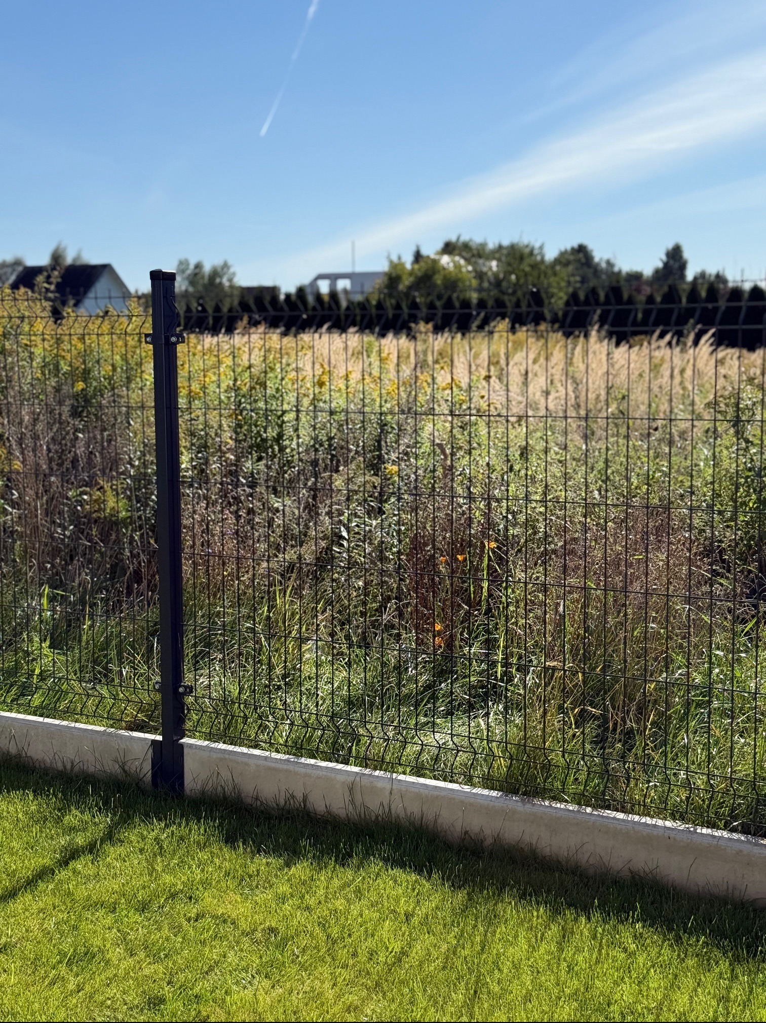 Ciemny panel ogrodzeniowy z podmurówką na tle bujnej roślinności i błękitnego nieba. Widoczny fragment trawnika. Słoneczny dzień.