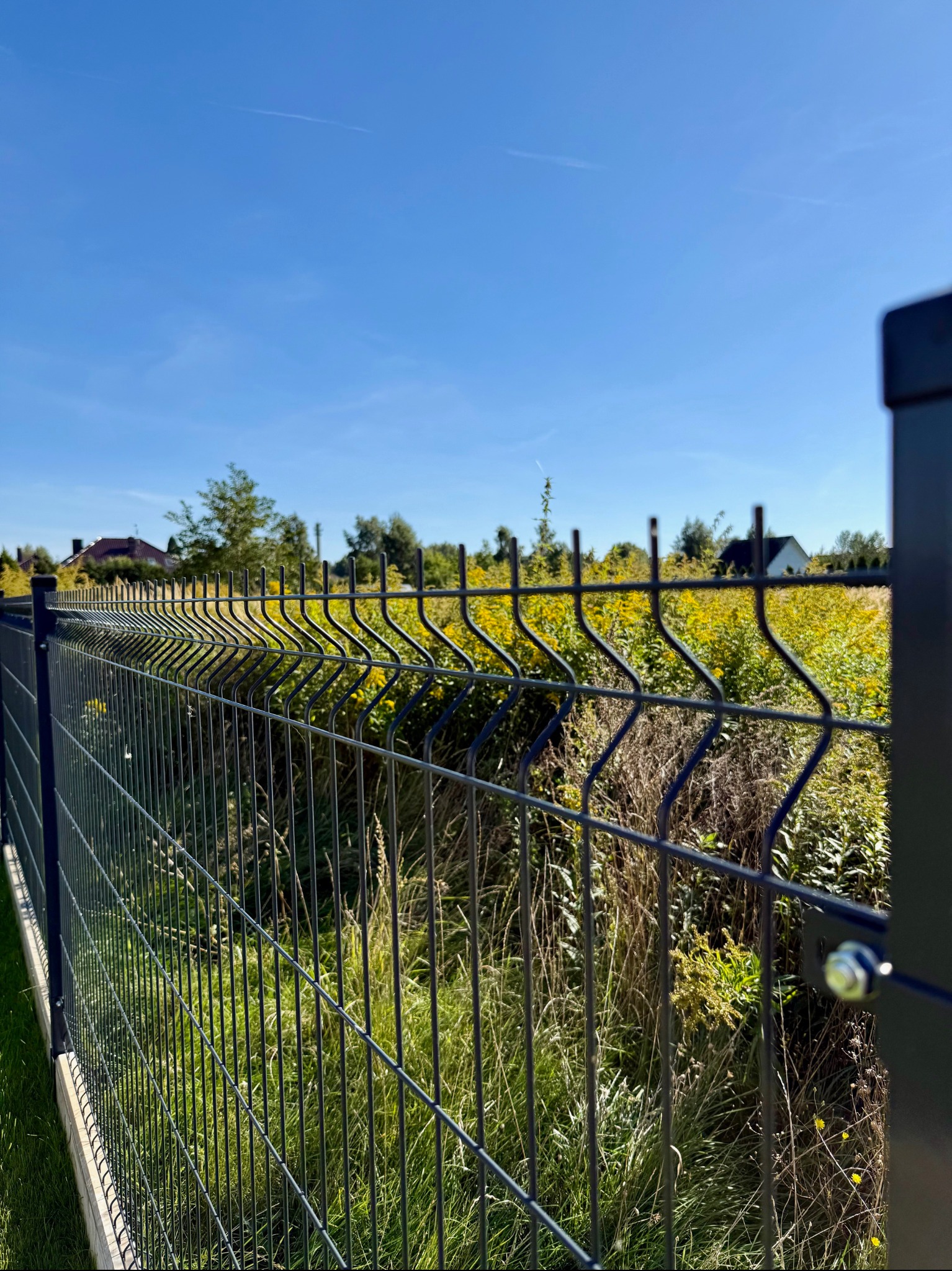 Nowoczesne ogrodzenie panelowe w kolorze grafitowym na tle zieleni i błękitnego nieba. Widoczne detale montażowe i solidna konstrukcja. Ochrona posesji z dbałością o estetykę.