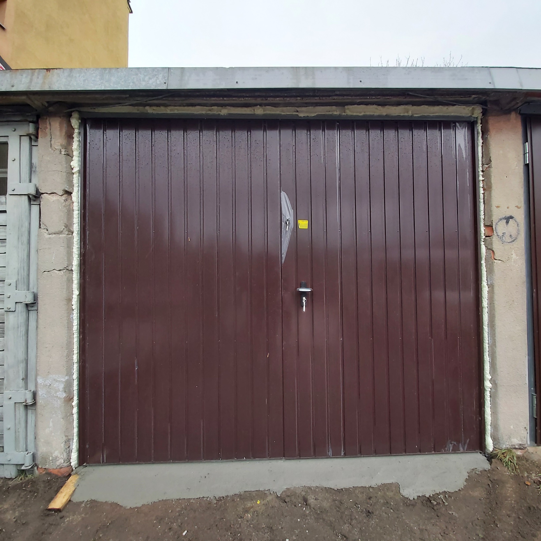 Brązowa brama garażowa segmentowa po montażu, uszczelniona pianką montażową, z widocznymi śladami po folii ochronnej i żółtą naklejką informacyjną.