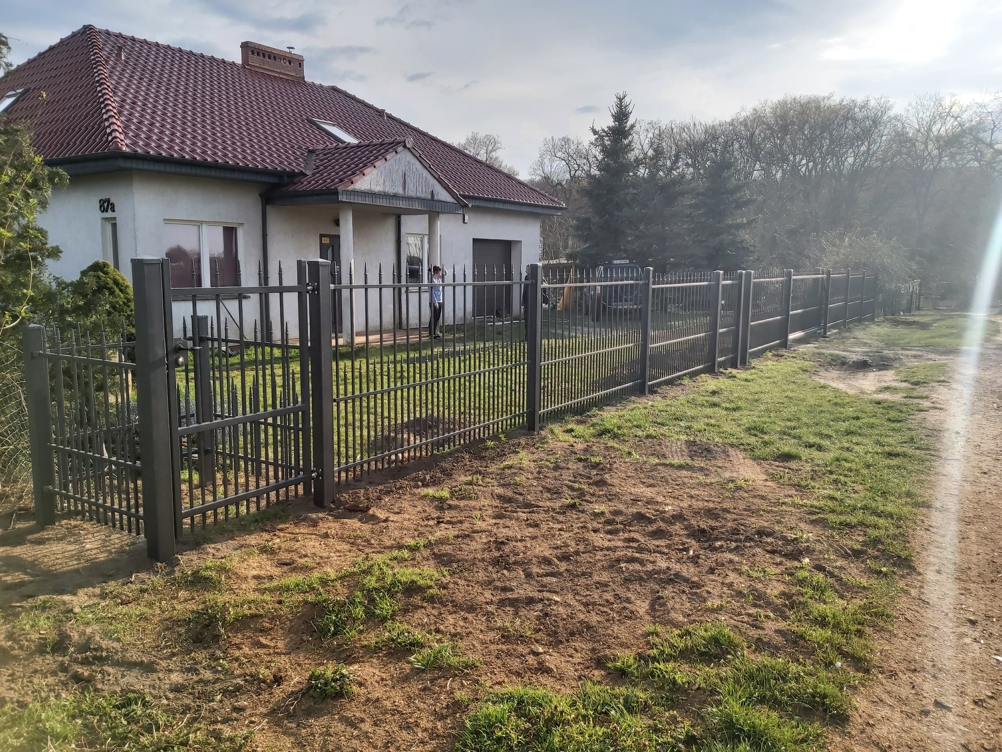 Nowoczesne, grafitowe ogrodzenie z metalowych prętów otaczające dom z czerwoną dachówką; w tle widać drzewa i fragment drogi.