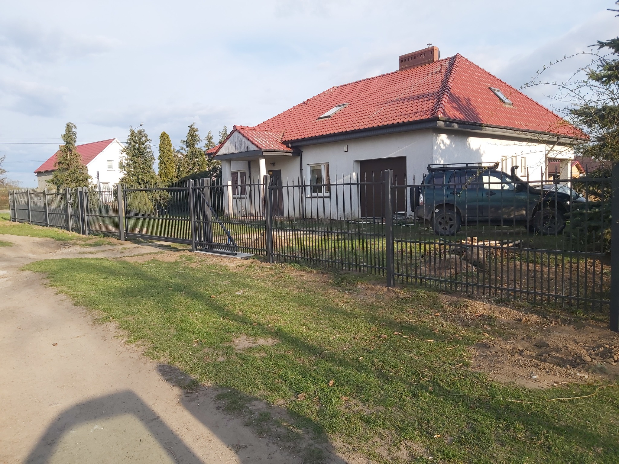 Nowoczesne, czarne ogrodzenie z metalowych prętów ogradza posesję z białym domem z czerwoną dachówką, na podwórku stoi terenowy samochód.