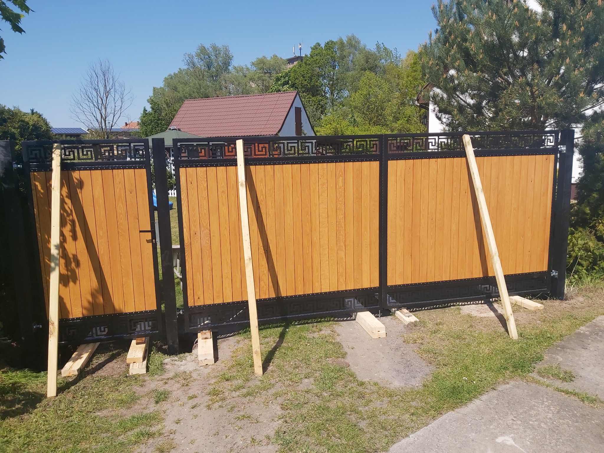 Trzyczęściowa brama wjazdowa z drewnianymi panelami i czarną, metalową ramą w trakcie montażu, podparta drewnianymi belkami, na tle trawnika i zabudowań.