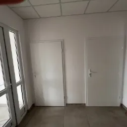 Wnętrze z dwoma białymi, zamontowanymi drzwiami i częściowo widocznym oknem balkonowym z białymi ramami, szara podłoga, widoczne kable i skrzynka narzędziowa w tle.