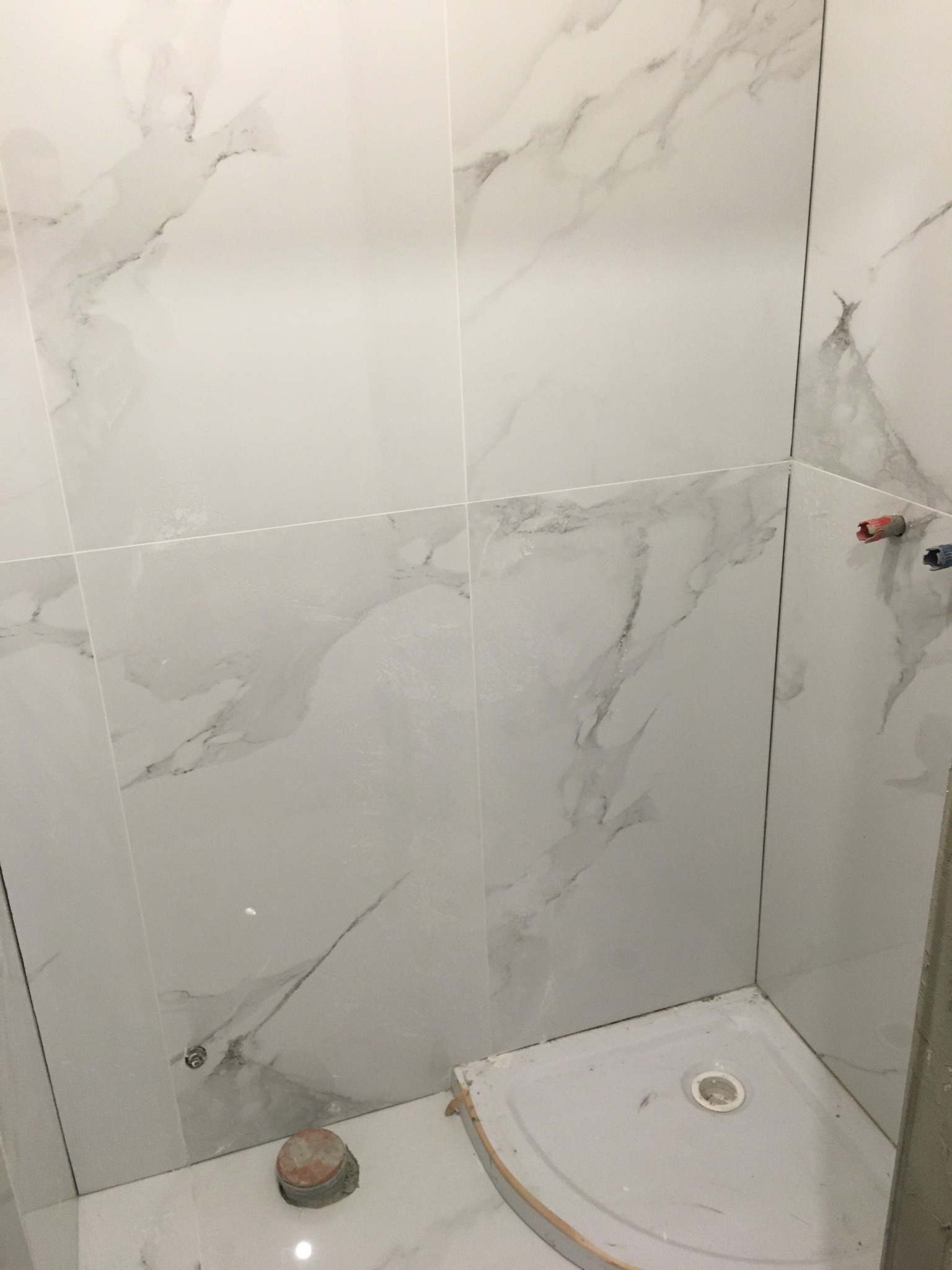 Ściana wyłożona jasnymi, marmurkowymi płytkami w kabinie prysznicowej, widoczne wystające rury z zaślepkami i odpływ w brodziku.