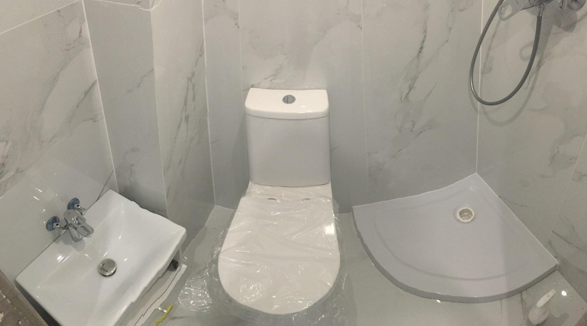 Nowa, biała łazienka z marmurowymi płytkami, umywalką, toaletą z folią ochronną i narożnym brodzikiem.