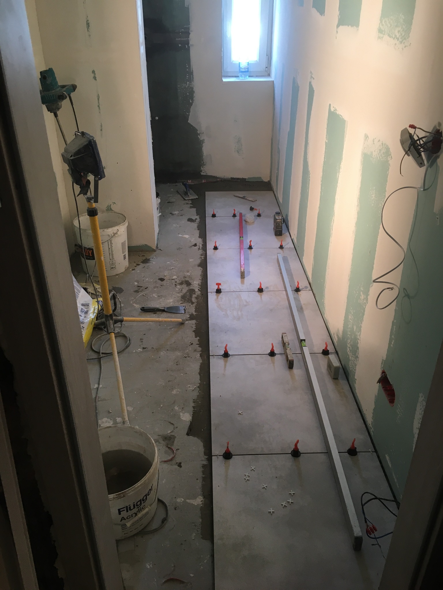Układanie szarych płytek podłogowych w trakcie remontu łazienki, widoczne poziomice, krzyżyki dystansowe i wiadro z wodą marki Flugger Acrylic.