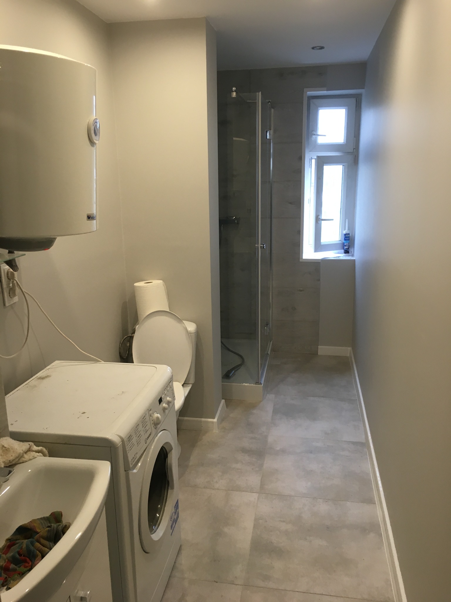 Wnętrze wyremontowanej łazienki z kabiną prysznicową, pralką obok umywalki, bojlerem na ścianie i oknem z silikonem na parapecie.