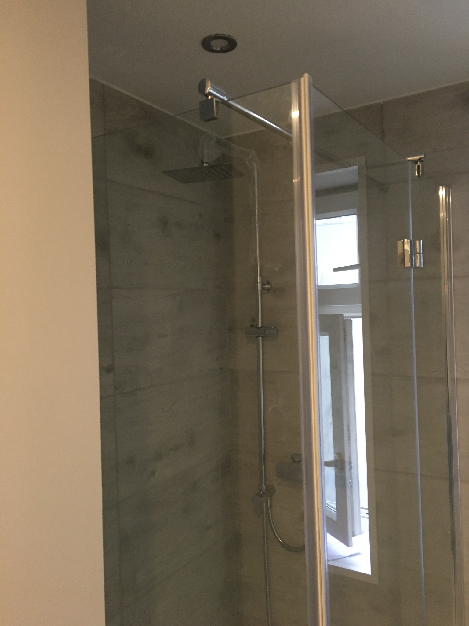 Nowoczesna kabina prysznicowa z transparentnego szkła z widocznym oknem uchylnym w tle, wykończona płytkami imitującymi drewno.