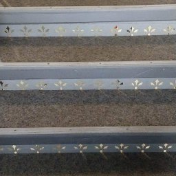 Dag-Bud S.C. - Schody z szarego dywanu, z pomalowanymi na szaro stopnicami ozdobionymi wyciętymi wzorami w kształcie stylizowanych kwiatów.