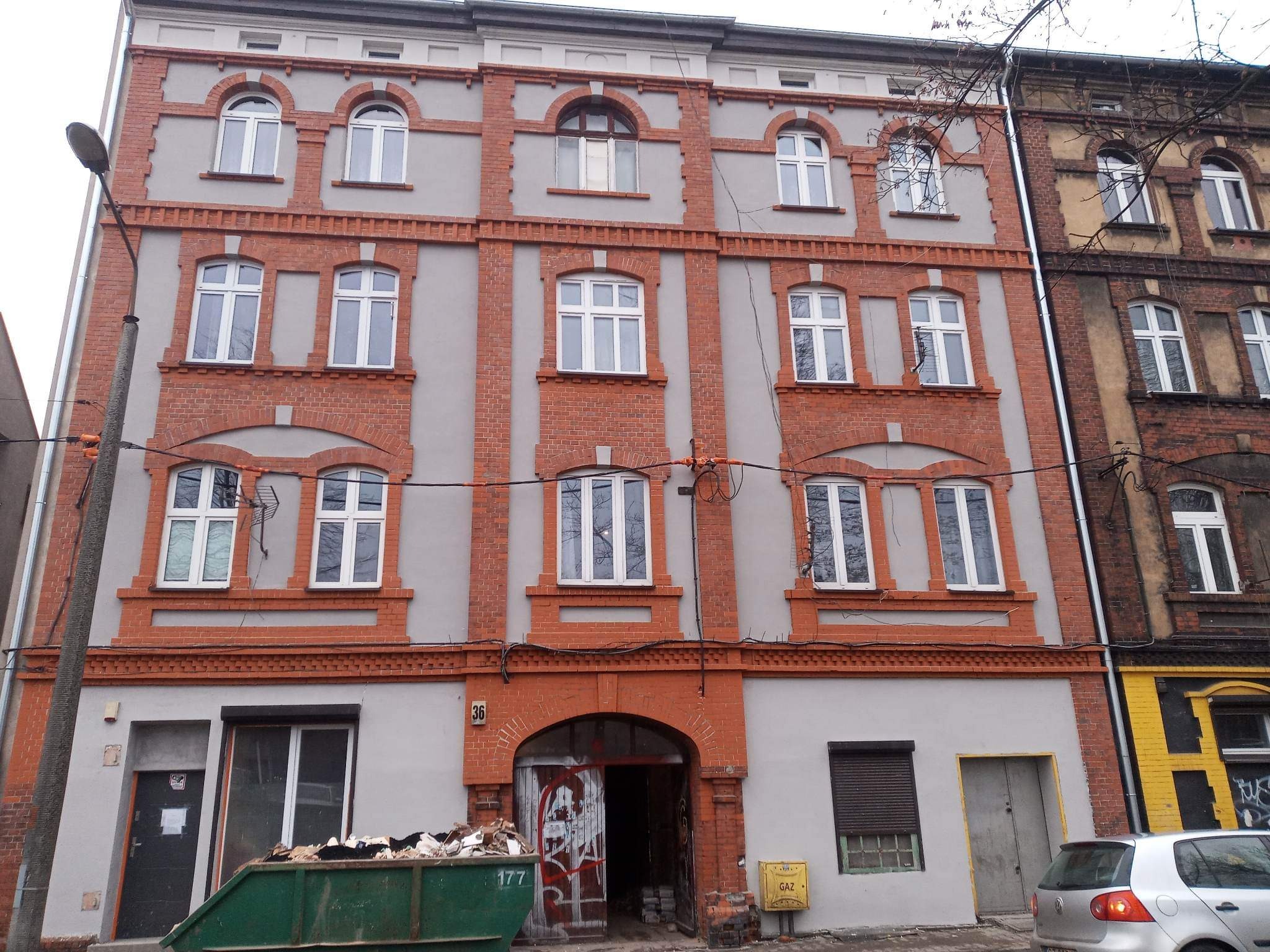 Fasada kamienicy po częściowym odnowieniu, z widocznym kontenerem na gruz przed wejściem i fragmentem sąsiedniej, nieodnowionej kamienicy.