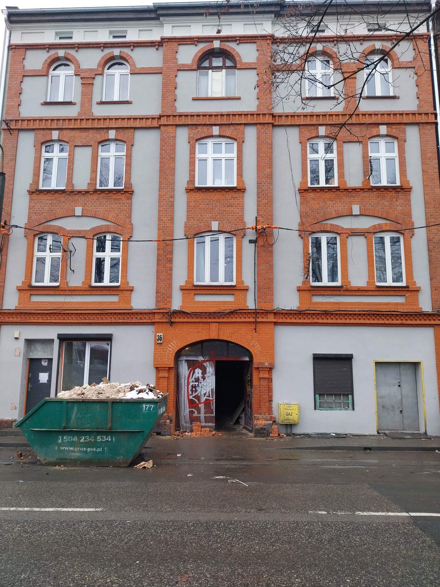Budynek z ceglaną elewacją w trakcie remontu, widoczny kontener ze gruzem przed wejściem.