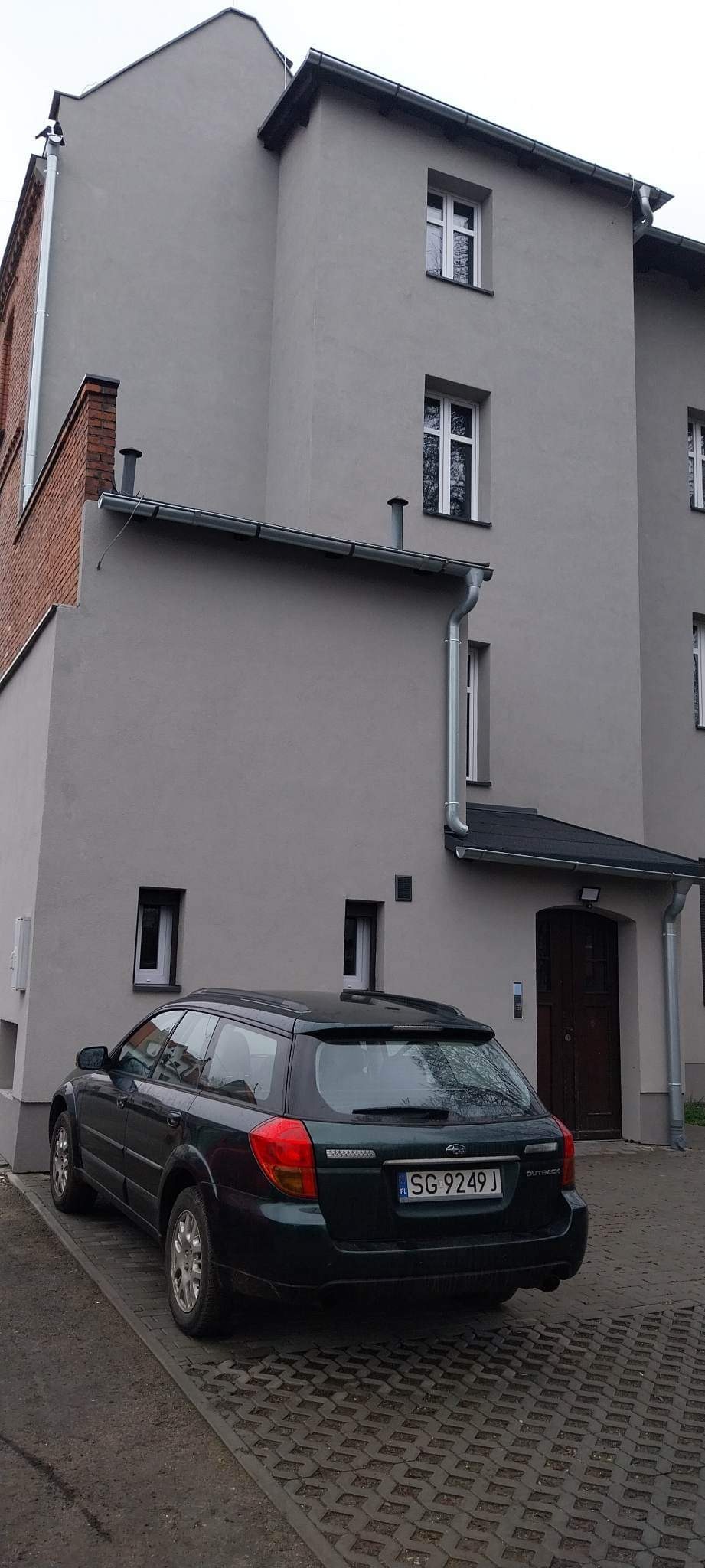 Szara elewacja budynku wielopiętrowego z nowymi oknami i rynnami, zaparkowany ciemnozielony samochód osobowy na ażurowej kostce brukowej.