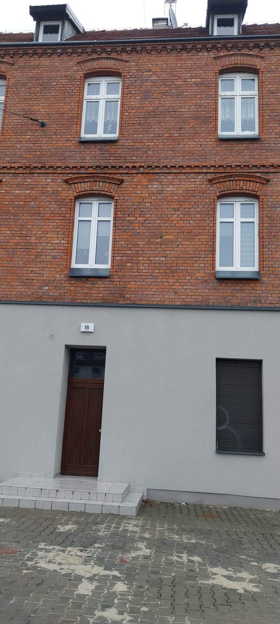 Odnowiona fasada budynku z czerwonej cegły z nową, gładką, szarą elewacją poniżej, widoczne białe okna z dekoracyjnymi łukami i ciemne, drewniane drzwi wejściowe z numerem '18' nad nimi.