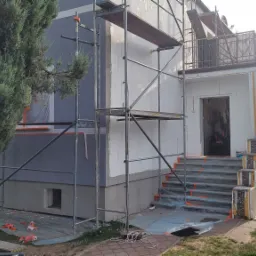 Dom jednorodzinny w trakcie prac elewacyjnych: ocieplanie styropianem, częściowo pomalowany na szaro, widoczne rusztowania, zabezpieczone schody wejściowe folią, obok rośnie iglak.