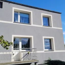 Dwukondygnacyjny dom z szarą elewacją, białymi obramowaniami okien i poziomymi pasami, widoczny z perspektywy balkonu z metalową balustradą, w tle błękitne niebo z chmurami.