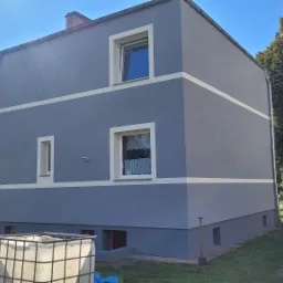 Dwupiętrowy dom po tynkowaniu i malowaniu elewacji na szaro z białymi pasami, widoczne wiadro z farbą i szpadel przy ścianie.