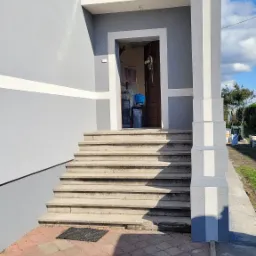 Wejście do domu po remoncie: świeżo pomalowana elewacja w odcieniach szarości z białymi akcentami, schody wyłożone kamieniem prowadzące do otwartych drzwi.