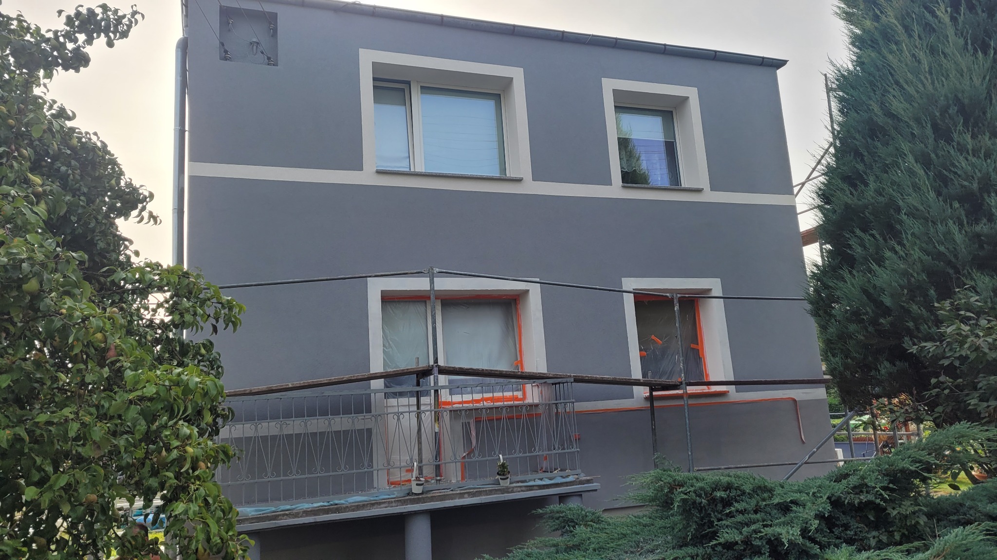 Dwukondygnacyjny dom w trakcie malowania elewacji na szaro, okna zabezpieczone folią, widoczne rusztowania i zieleń drzew.
