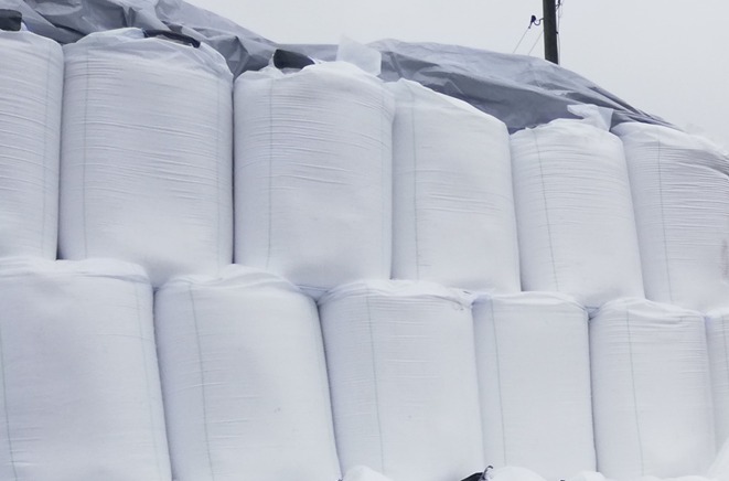 Stos białych, worków z nawozem lub zbożem, zabezpieczonych szarą plandeką, składowanych na zewnątrz w pochmurny dzień.