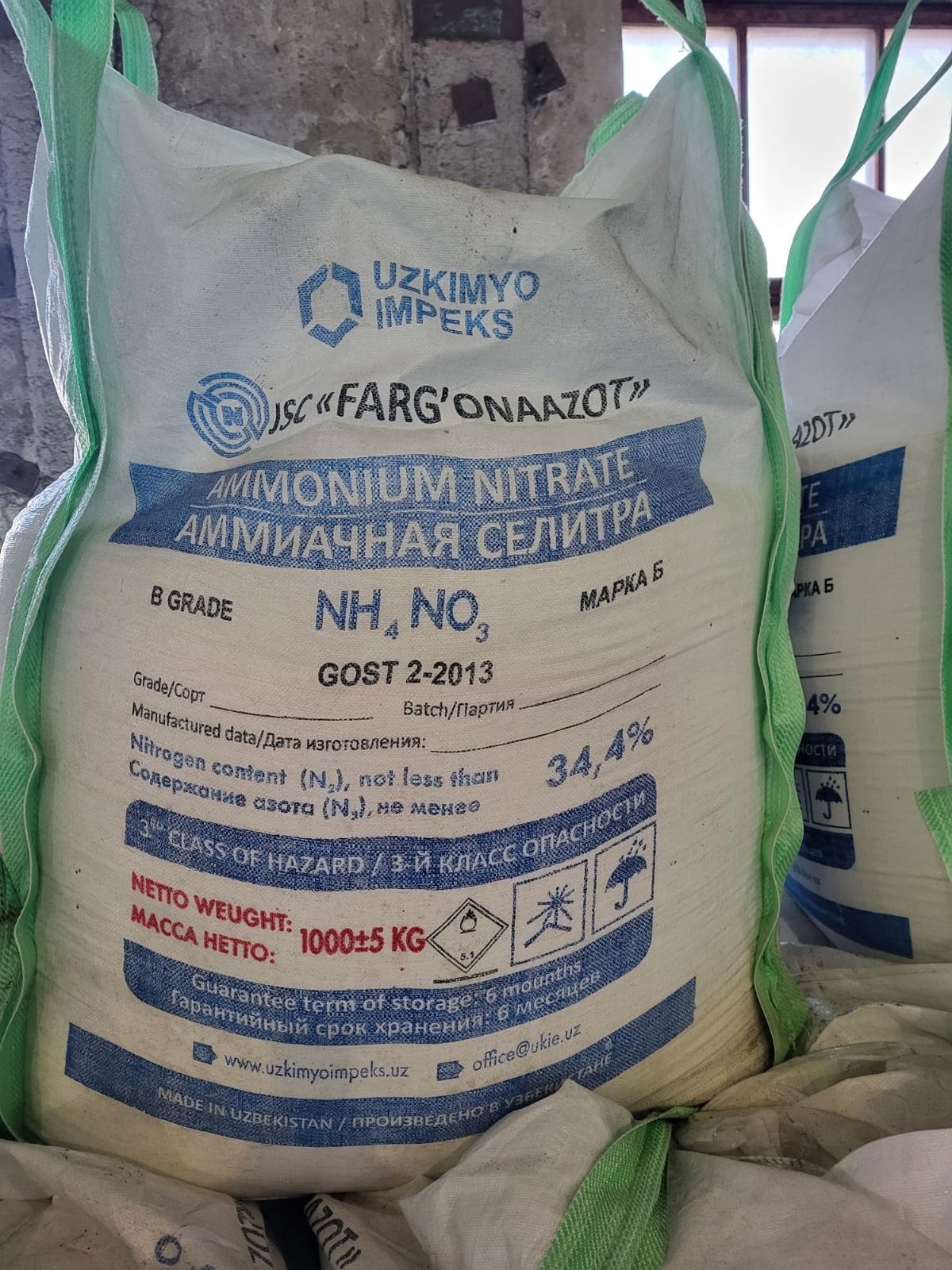 Wielki, biały worek z nawozem azotowym (saletra amonowa) z niebieskimi napisami i oznaczeniami, w magazynie, widoczne inne worki w tle.