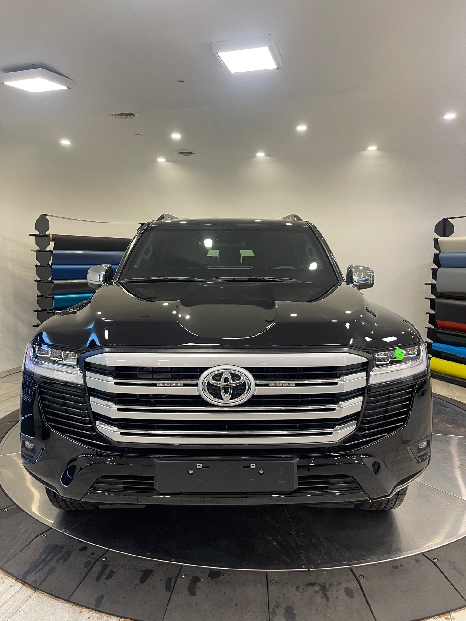 Czarny samochód Toyota Land Cruiser na obrotowej platformie w jasnym salonie, z widocznymi rolkami folii do oklejania pojazdów w tle.