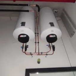 Dwa białe podgrzewacze wody zamontowane na ścianie, połączone rurami miedzianymi z zaworami, widoczne rury instalacyjne pod sufitem i fragment okna.