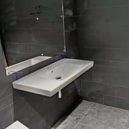 Bartłomiej Fita - Nowoczesna łazienka w trakcie remontu: biała, wisząca umywalka z chromowanym syfonem na tle ciemnych, prostokątnych płytek ściennych i podłogowych, widoczne ślady prac remontowych na podłodze...