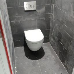 Bartłomiej Fita - Nowoczesna toaleta podwieszana z chromowanym przyciskiem spłukującym, osadzona na tle grafitowych płytek imitujących kamień w wykończonej łazience.