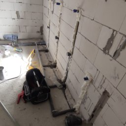 Bartłomiej Fita - Ściana z betonu komórkowego z wyciętymi bruzdami wypełnionymi pianką montażową wokół rur instalacji wodnej, torba narzędziowa leżąca na podłodze.