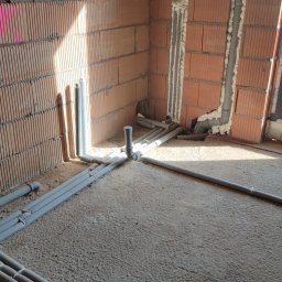Bartłomiej Fita - Surowe wnętrze z ceglanymi ścianami i szarymi rurami instalacji wodnej lub grzewczej ułożonymi na podłodze w trakcie montażu.