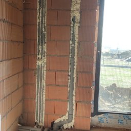 Bartłomiej Fita - Wewnętrzne ściany z cegły z bruzdami wypełnionymi pianką montażową wokół szarych rur kanalizacyjnych, widoczne rury na podłodze w surowym stanie, okno z profilem VEKA.