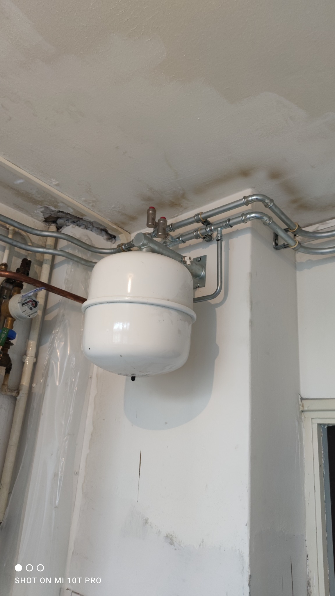 Biały zbiornik wyrównawczy c.o. zamontowany na ścianie, widoczne rury instalacji grzewczej z zaworami, oraz licznik wody w tle, sufit z widocznymi śladami wilgoci.