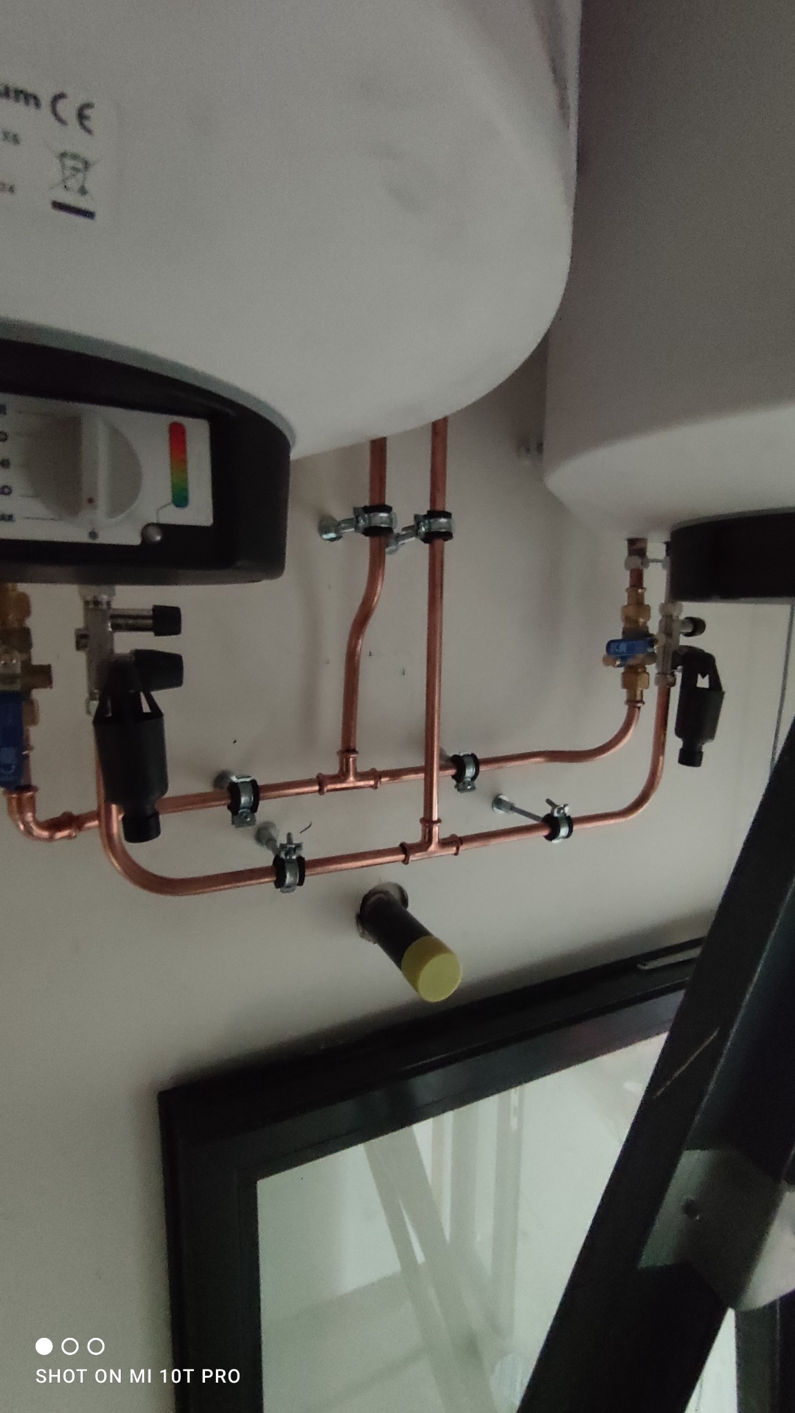 Kompleksowa instalacja dwóch podgrzewaczy wody z miedzianymi rurami, zaworami i filtrami, zamontowana na ścianie, widoczna fragment ramy okiennej.