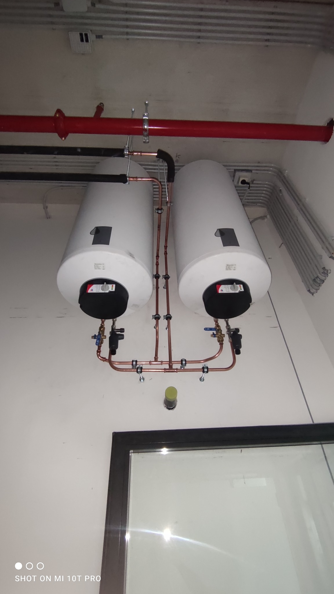 Dwa białe podgrzewacze wody zamontowane na ścianie, połączone rurami miedzianymi z zaworami, widoczne rury instalacyjne pod sufitem i fragment okna.