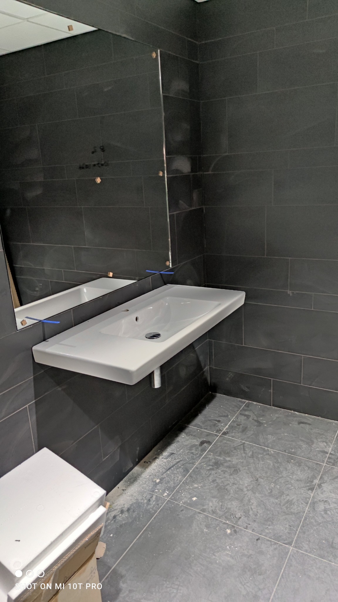 Nowoczesna łazienka w trakcie remontu: biała, wisząca umywalka z chromowanym syfonem na tle ciemnych, prostokątnych płytek ściennych i podłogowych, widoczne ślady prac remontowych na podłodze...