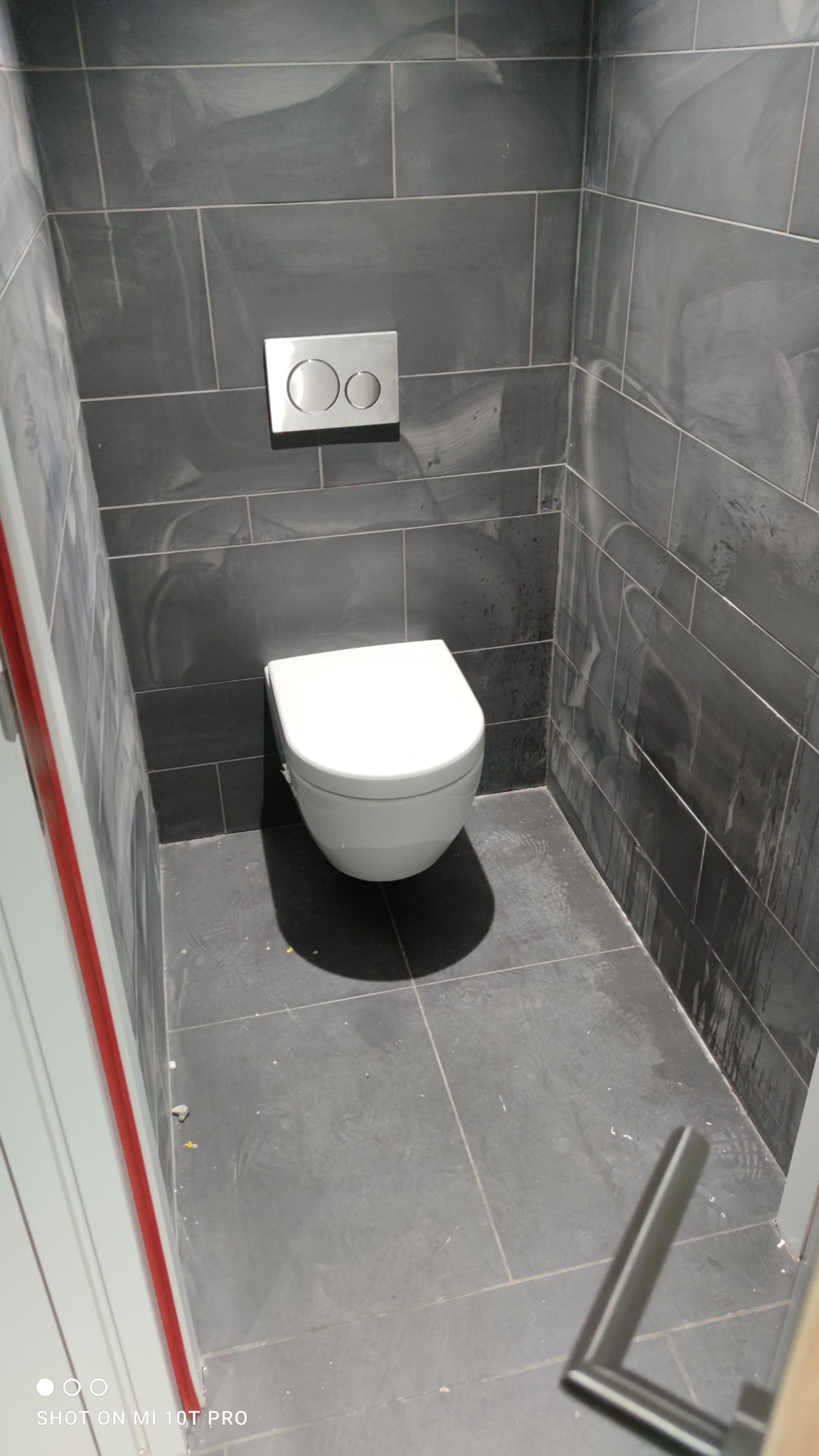 Nowoczesna toaleta podwieszana z chromowanym przyciskiem spłukującym, osadzona na tle grafitowych płytek imitujących kamień w wykończonej łazience.