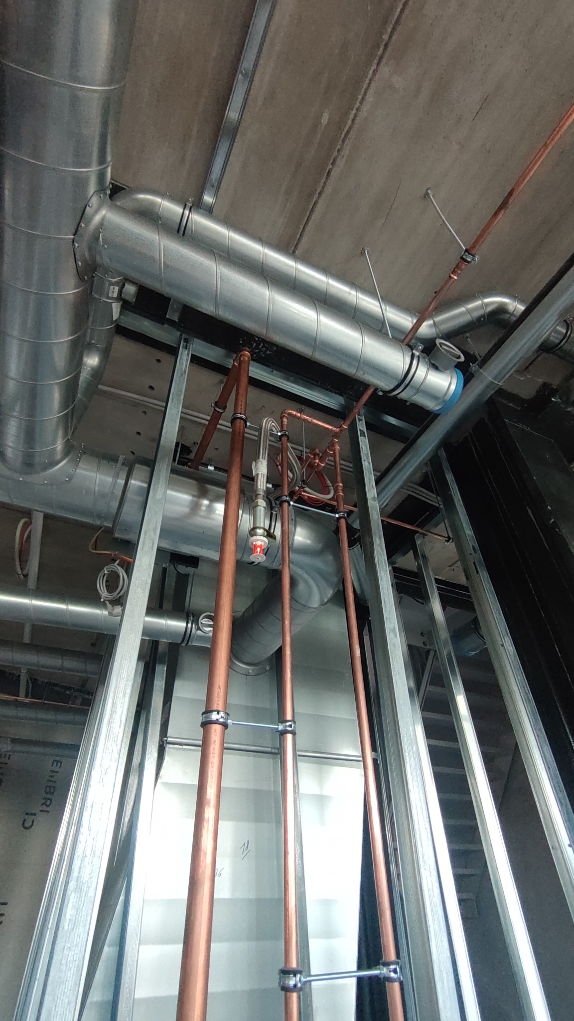 Widok z dołu na instalację wentylacyjną z rurami miedzianymi i elementami mocującymi pod sufitem, w trakcie montażu, widoczne profile konstrukcyjne.