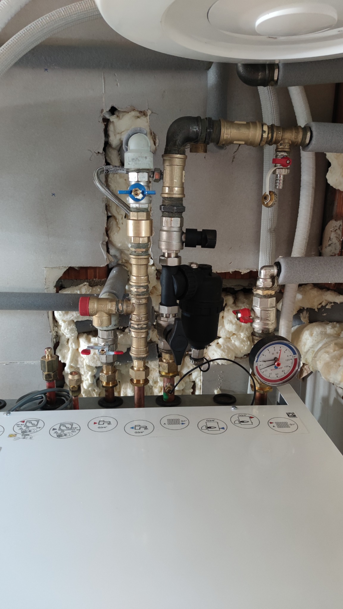 Przyłącza instalacji grzewczej z widocznymi zaworami, manometrem i izolacją piankową na ścianie, instalacja częściowo ukryta za płytą kotła z piktogramami.