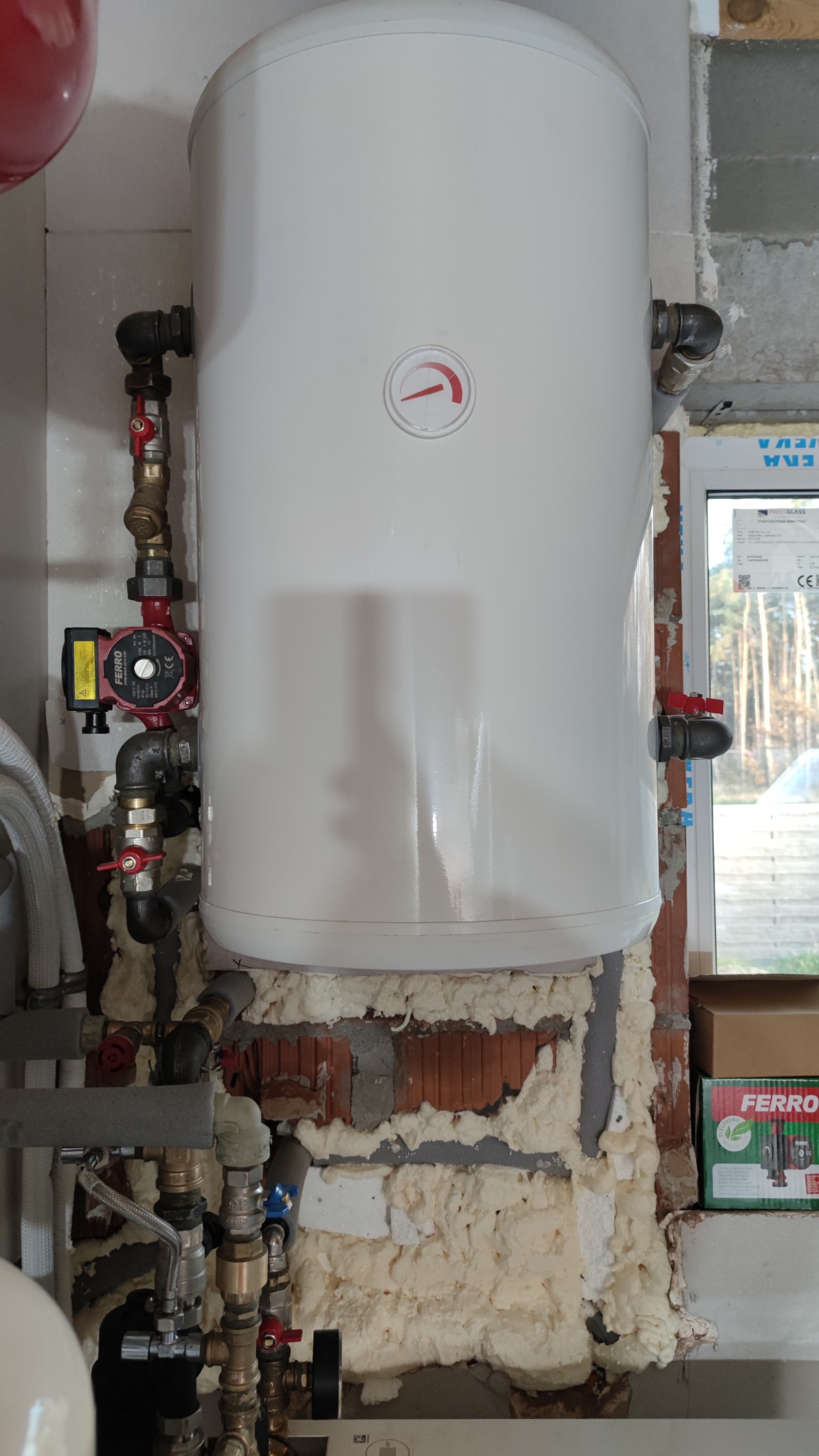Biały bojler z czerwonym wskaźnikiem, zamontowany przy oknie, z widocznymi rurami, zaworami i pompą obiegową, otoczony pianką montażową na surowej ścianie z cegieł.