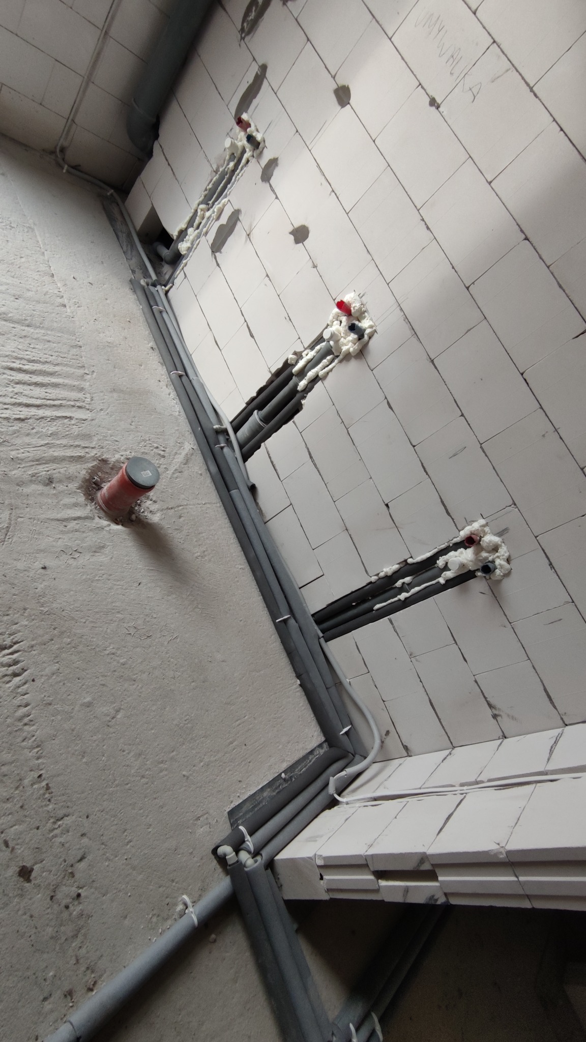 Szare rury instalacyjne prowadzone wzdłuż ściany z białych bloczków, zabezpieczone pianką montażową w miejscach przejść przez ścianę; widoczna również rura wystająca z podłogi.